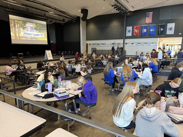 a candid photo of several students sitting around several cafeteria tables playing games, watching a movie and coloring  