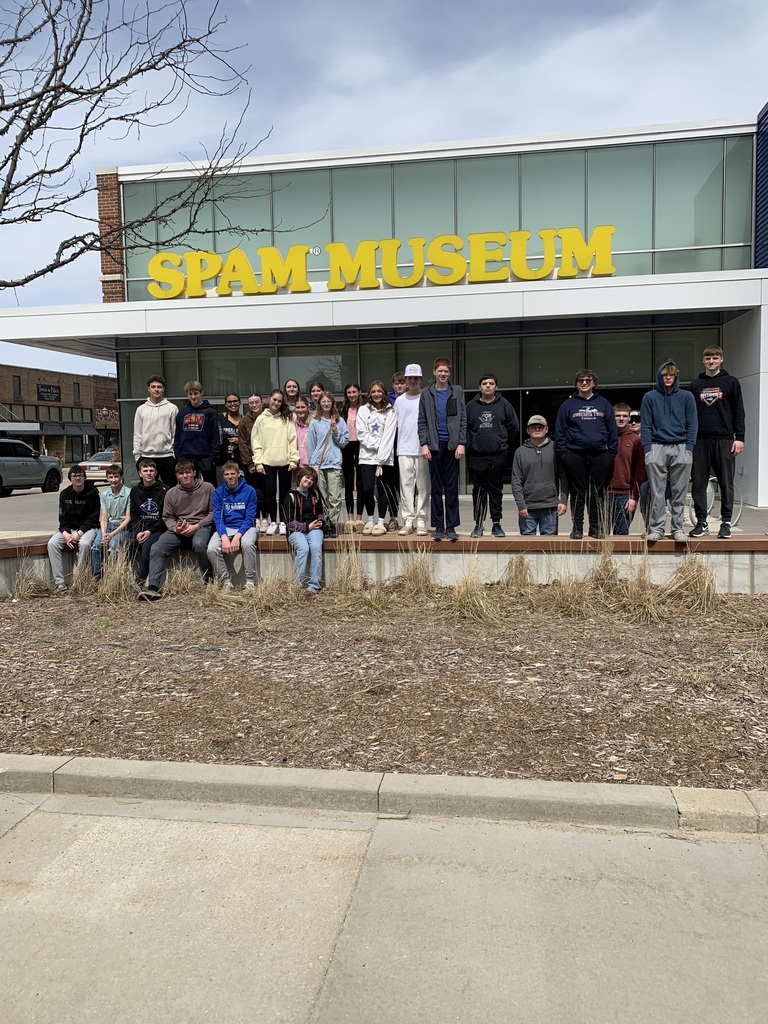 Students posing in front of the Spam Museum in a line.  Some students are sitting on the concrete ledge in front of the students who are standing. 
