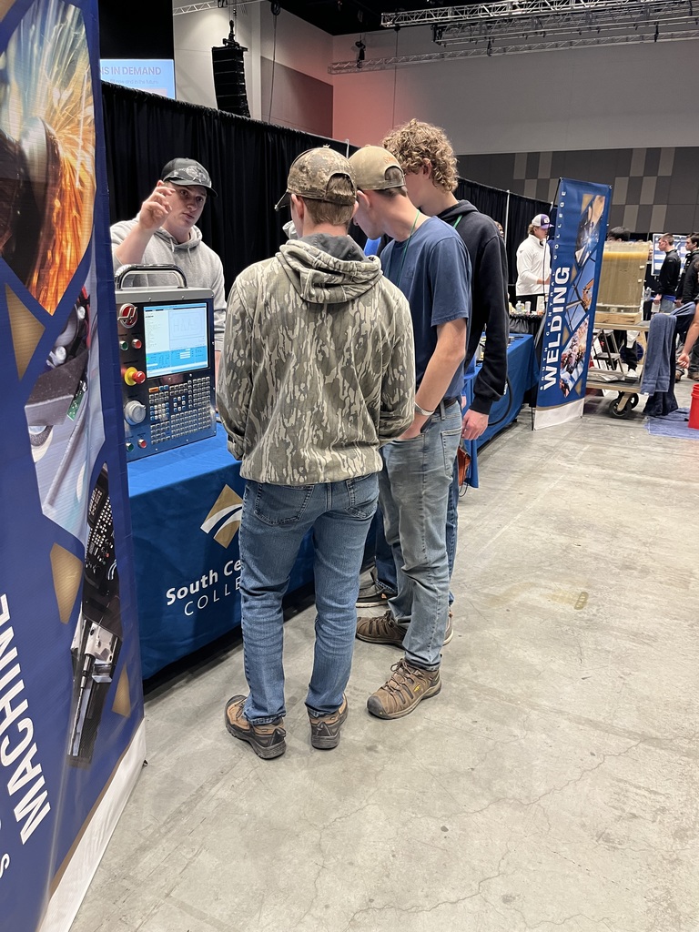 Students standing in front of a table with a  rep from South Central College talking to the small group of three boys.  
