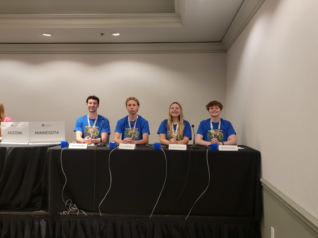 students on stage, sitting at a table, competing wearing blue shirts and medals with their names in front of them on the table.  