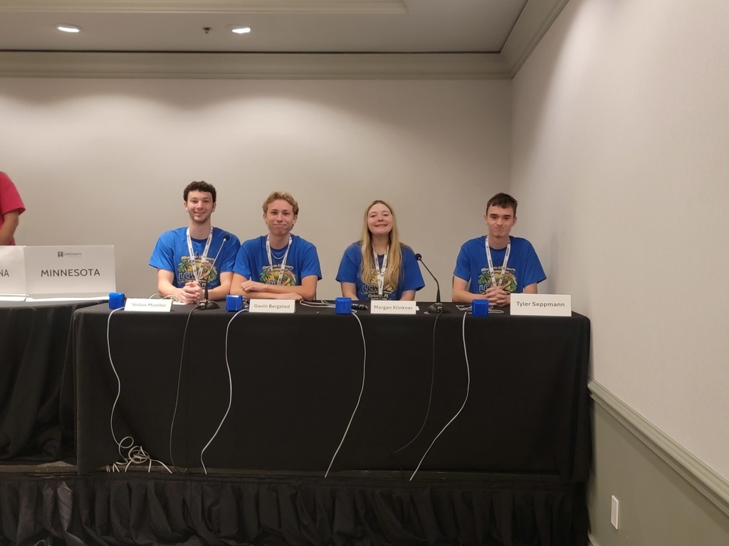 students on stage, sitting at a table, competing wearing blue shirts and medals with their names in front of them on the table.  