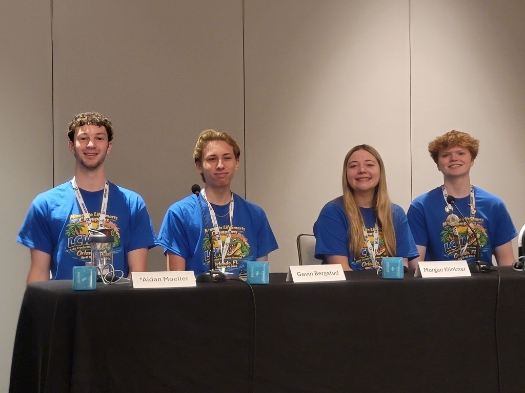 students on stage, sitting at a table, competing wearing blue shirts and medals with their names in front of them on the table.  