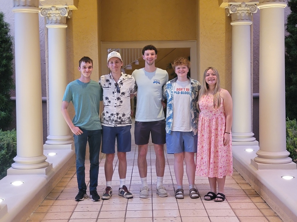 Students posing in between white pillars with lush green around them on a tile floor. 