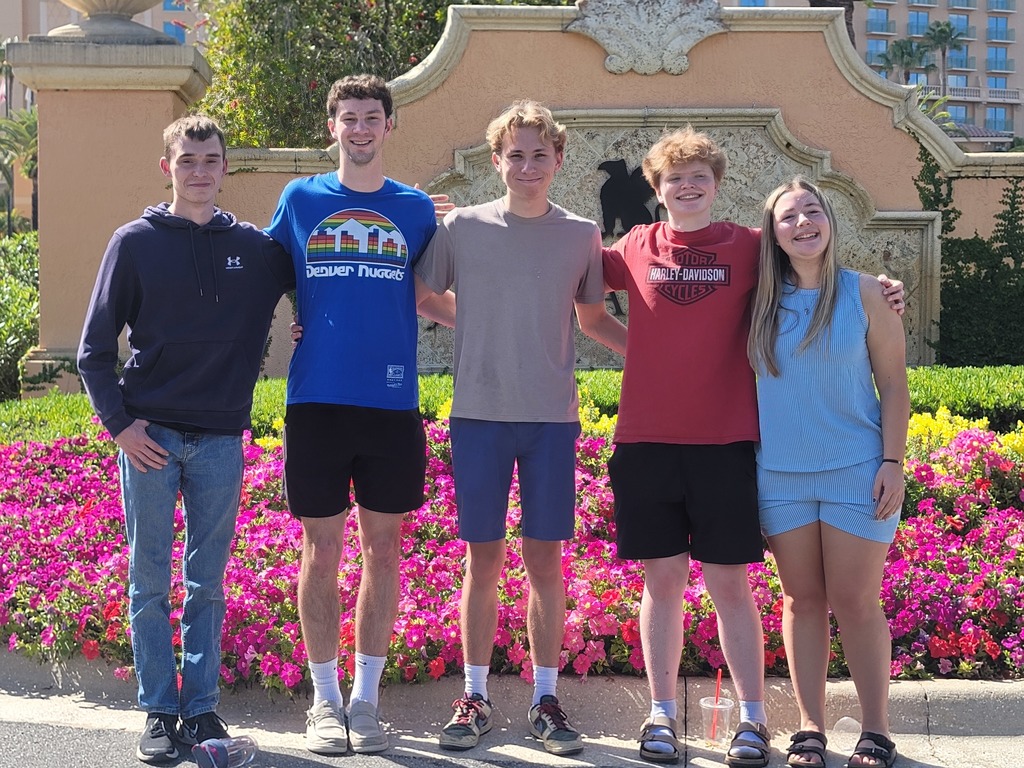 Students posing in front of a flower garden with pink flowers and green grass behind them.  They're smiling and looking at the camera. 