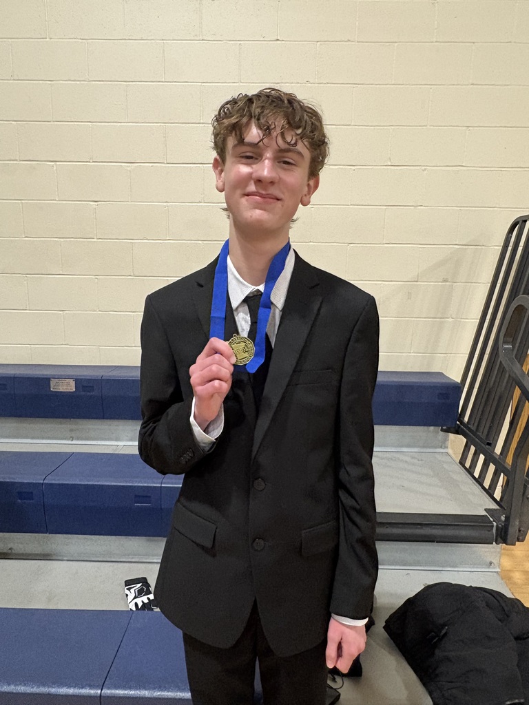 Ryan standing in the bleachers at a school with a white wall behind him, holding his blue medal to show he moved on to the state tournament.  