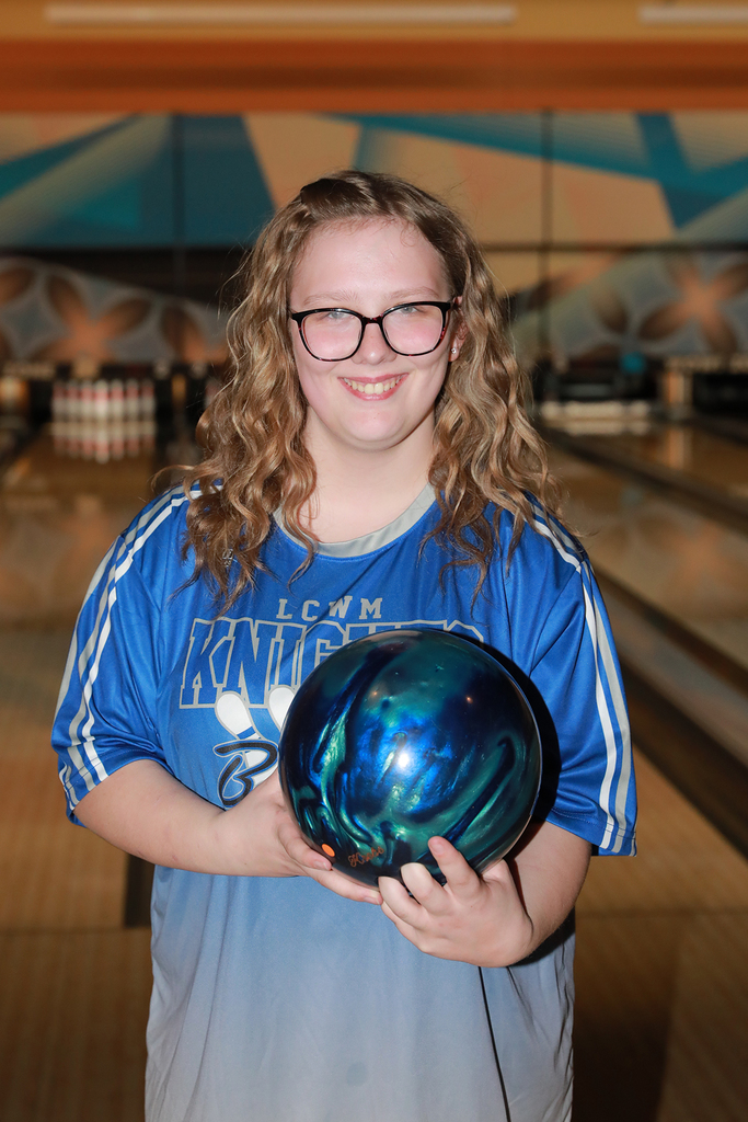 student smiling at the camera holding her bowling ball in front of the lanes.