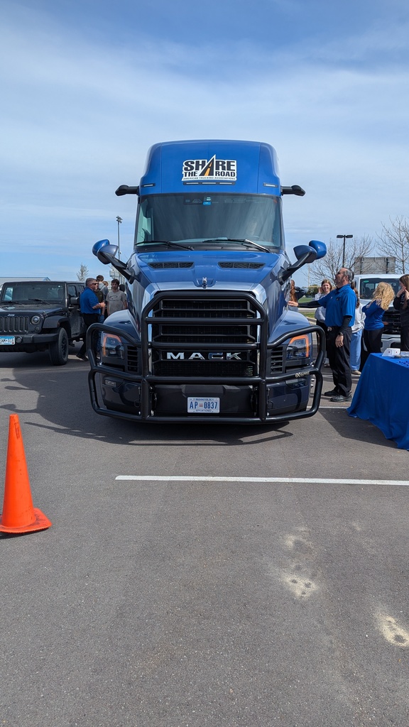 Straight on view from in front of the semi.  A student is sitting in the driver seat of a semi to experience the blindspots like a semi driver does while people stand on either side of the semi.  