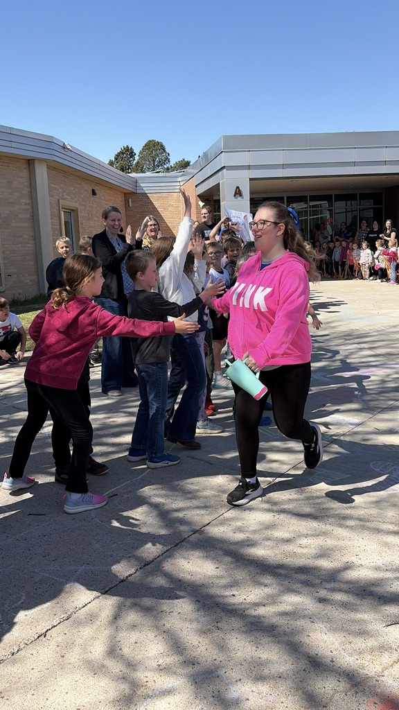 Carynn is walking out of the building high fiving a bunch of students standing outside of the school.  