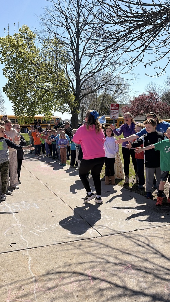 Carynn is walking out of the building high fiving a bunch of students standing outside of the school.  