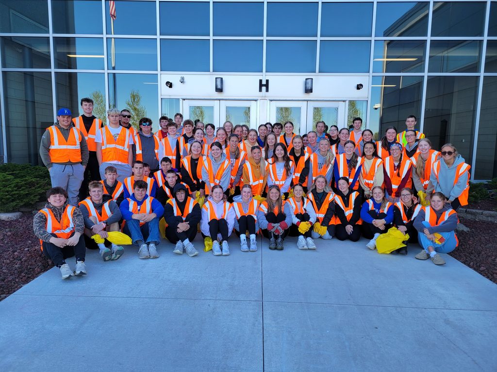 The whole group of NHS students standing and kneeling in front of the school doors with orange and yellow vests on.  