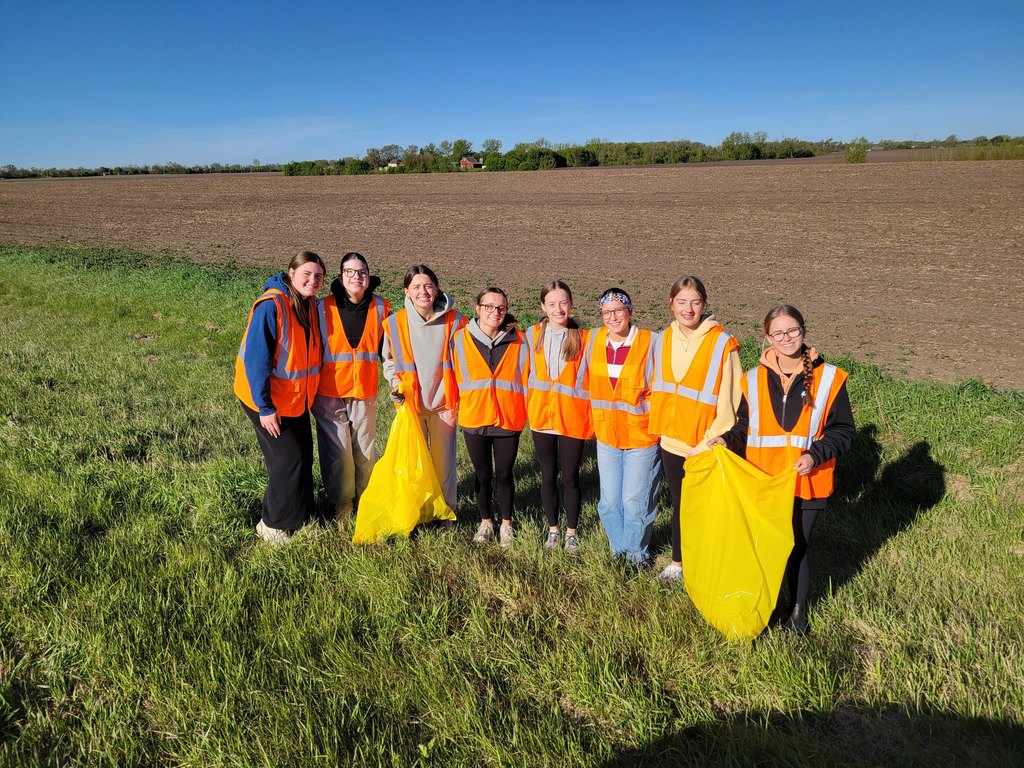 Students holding yellow garbage bags with orange vests on in the ditch picking up garbage.