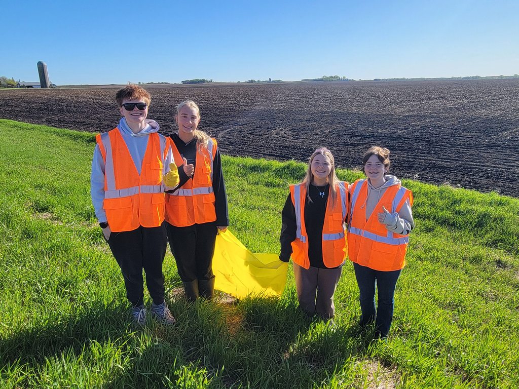 Students holding yellow garbage bags with orange vests on in the ditch picking up garbage.