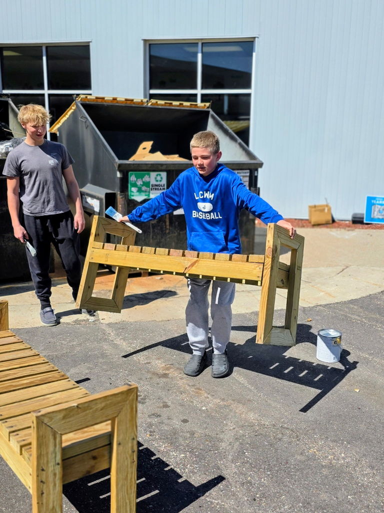 Student carrying a wooden bench with a paintbrush in one hand. 