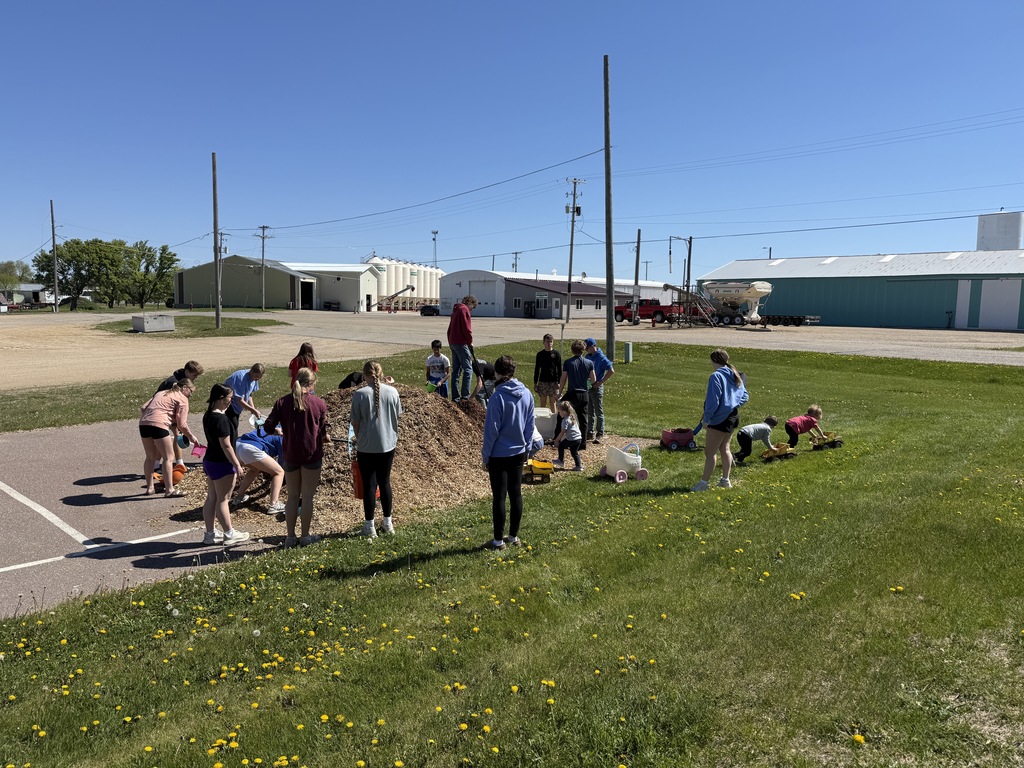 high school kids and little kids scooping mulch from a big pile and little kids pushing toy dump trucks with mulch in the back of them.  
