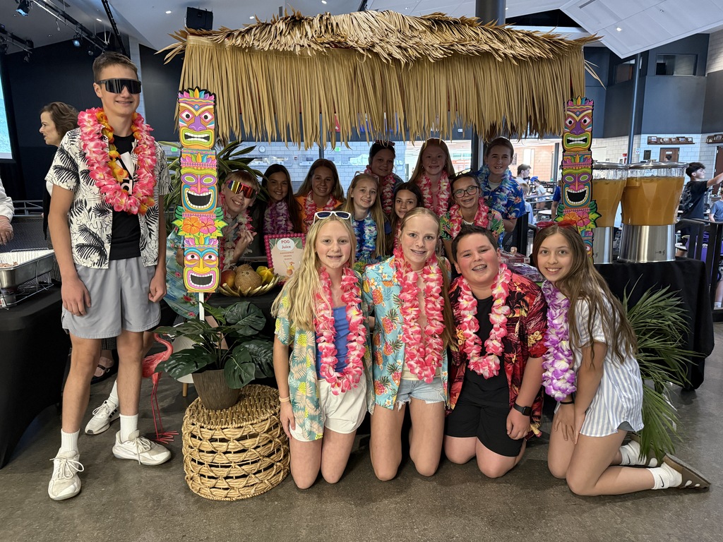 Middle School student council students with Hawaiian gear by a tiki tent with hawaiian  themed decor around it.