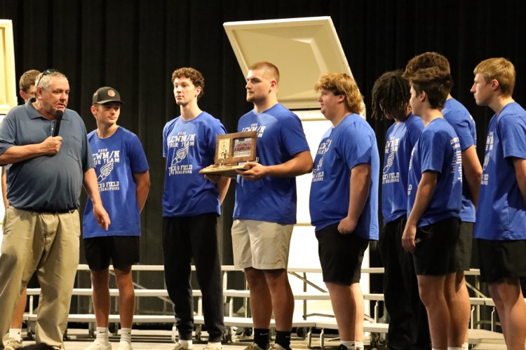 coach burns speaking into a mic while the team stands on the stage behind him. 