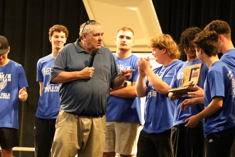 coach burns speaking into a mic while the team stands on the stage behind him. 