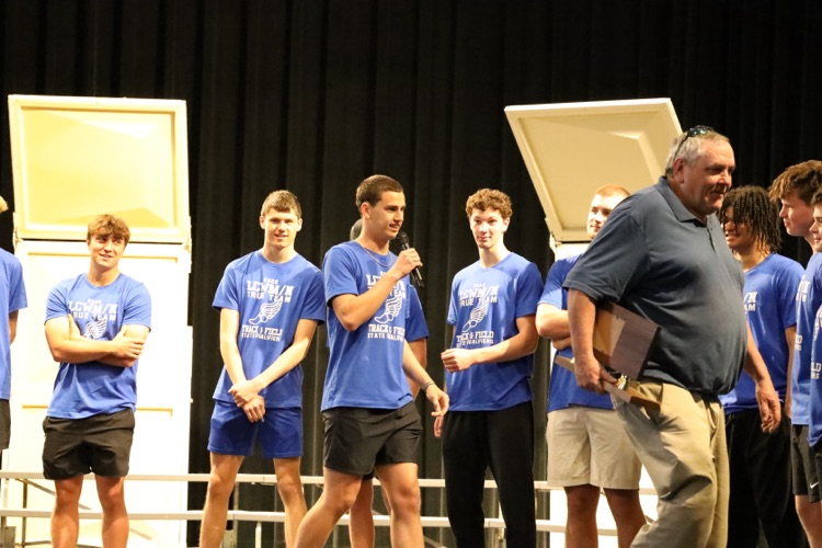Team member, Ryan masters speaking into a mic while the team stands on the stage behind him. 