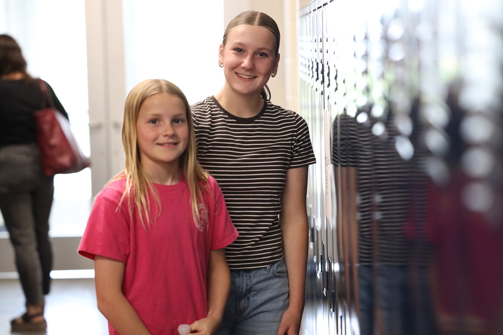 two students, one older, one younger standing by the lockers smiling at the camera. 