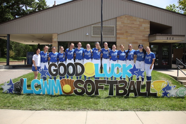 softball team lined up in front of the bank behind a sign that says good luck LCWM softball  