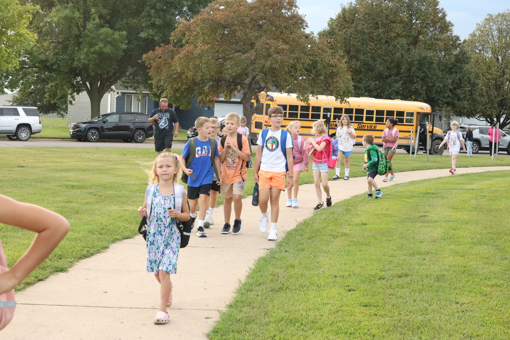 Students walking on a sidewalk up to school with a bus in the background.  