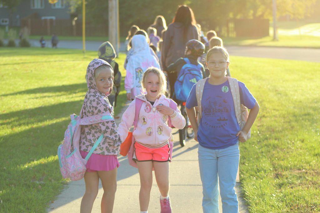Three students looking back at the camera with others in the background walking on the sidewalk towards the school.