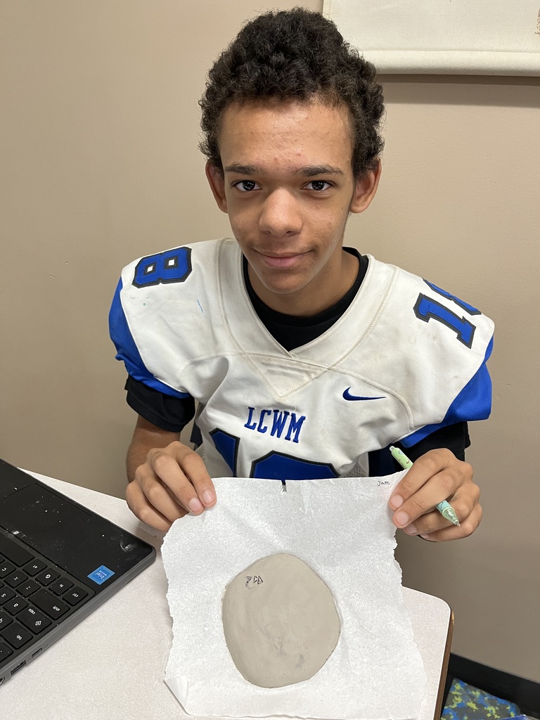 Student, looking and smiling at camera holding clay circle with cuneiform writing on it.