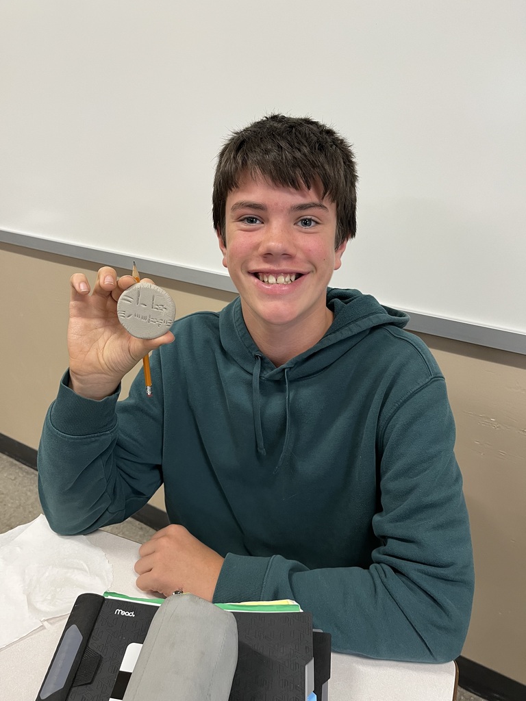 Student, looking and smiling at camera holding clay circle with cuneiform writing on it.