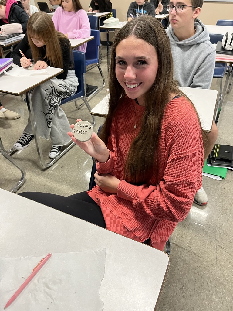 Student, looking and smiling at camera holding clay circle with cuneiform writing on it.