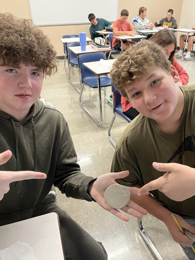 Students, looking and smiling at camera holding clay circle with cuneiform writing on it.