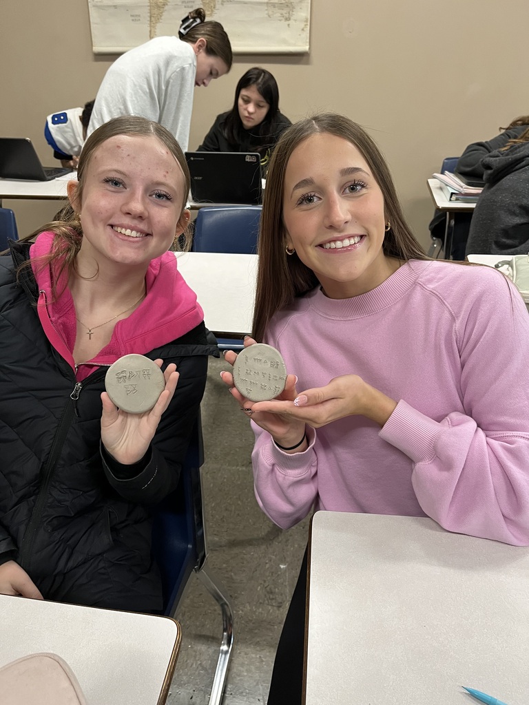 Student, looking and smiling at camera holding clay circle with cuneiform writing on it.