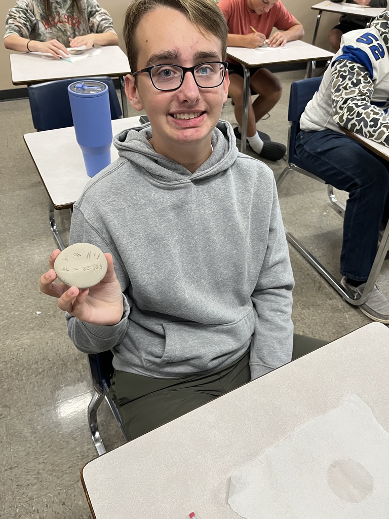 Student, looking and smiling at camera holding clay circle with cuneiform writing on it.