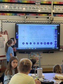 2 children in a classroom standing in front of graphs on a smart board