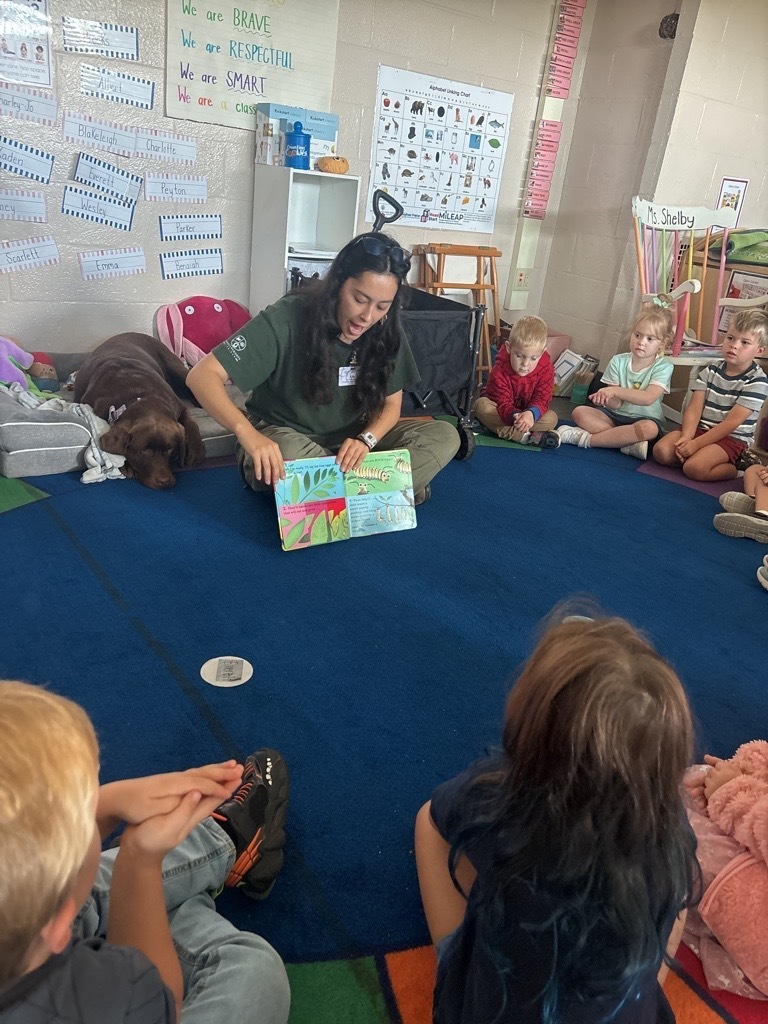 children sitting in a circle on a rug while a woman reads a book with a chocolate lab next to her