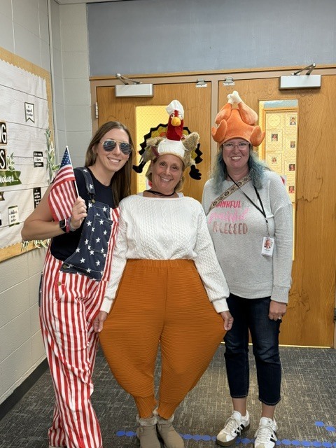 3 women posing together, 1 wearing american flag overalls, and 2 wearing turkey hats