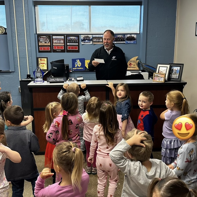 man standing behind his desk reading from a small piece of paper to a large group of children