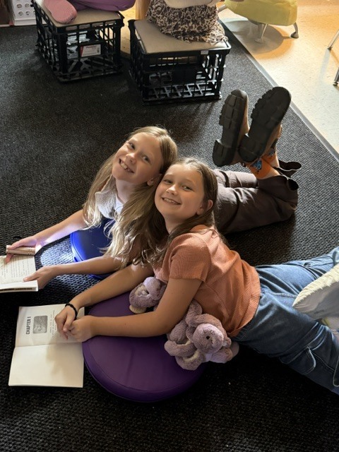 2 girls reading on a classroom floor with flashlights and stuffed animals