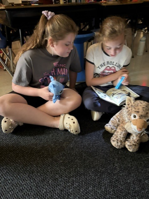 2 girls reading on a classroom floor with flashlights and stuffed animals