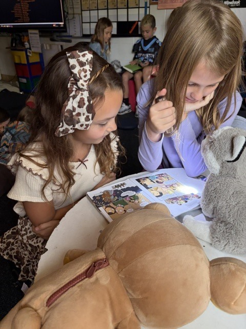 2 girls reading in a classroom with flashlights and stuffed animals