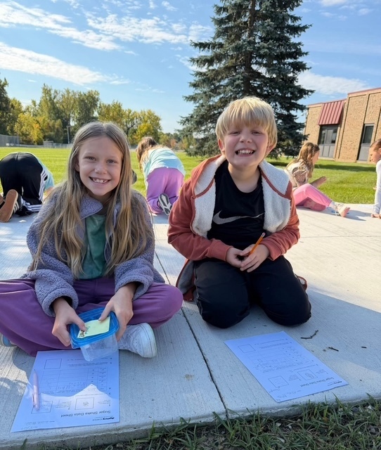 2 kids sitting on a sidewalk smiling