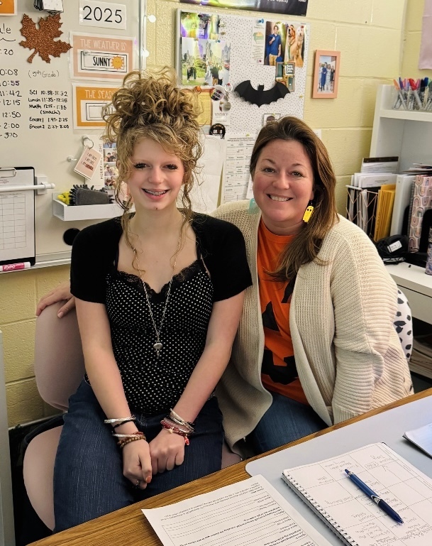 Two women sit together in a classroom at a desk smiling at the camera. Behind them is a wall with a whiteboard labeled "2025," weather posters, photos, a black bat cutout, and shelves with binders and supplies. On the desk are papers, a notebook, and a pen. One woman wears an orange top and banana-shaped earrings; the other wears a black polka-dot top with bracelets.