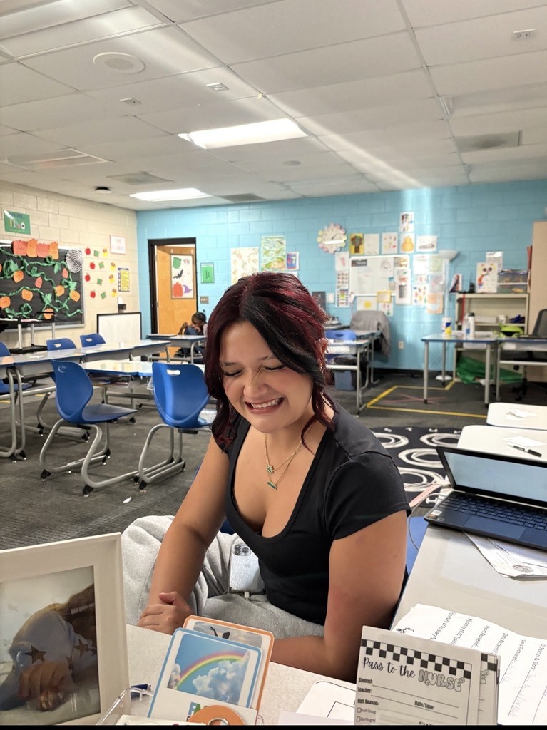 A girl with dark hair and a gray shirt sit at a teachers desk smiling. You can see the classroom behind her. 
