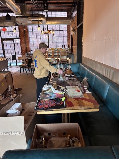 A person wearing a beige hoodie and dark pants looks at a display table with artwork, fabric, and decorative items inside a rustic café-like setting. The room features large grid-pane windows, exposed brick walls, warm hanging lights, an upholstered bench, and some boxes on the floor.