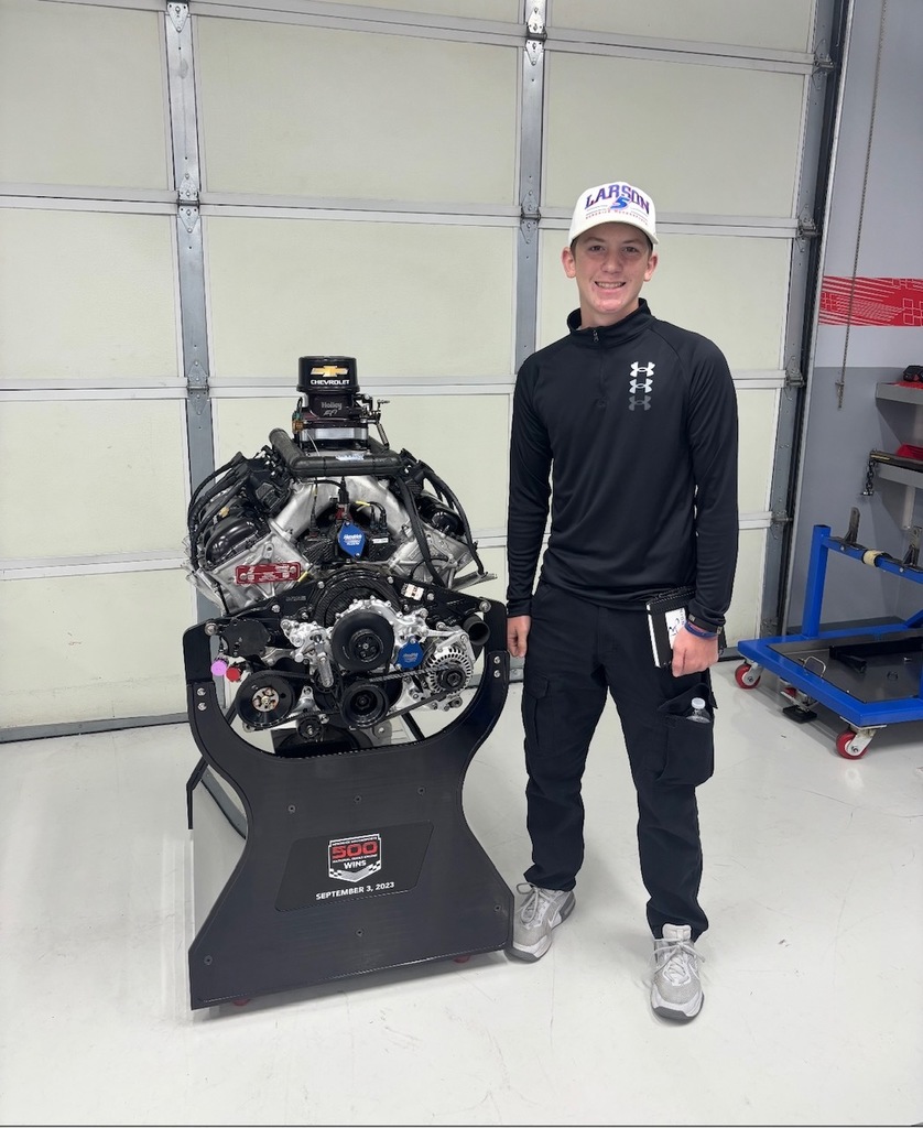 A young man smiles indoors next to a racing engine mounted on a stand. He wears a white cap labeled "LARSON," a black athletic jacket with Under Armour logo, black pants, and gray shoes. The background shows a garage or workshop with a large closed metal door and shelves. A sign on the engine stand reads: "500 WINS SEPTEMBER 3, 2023."
