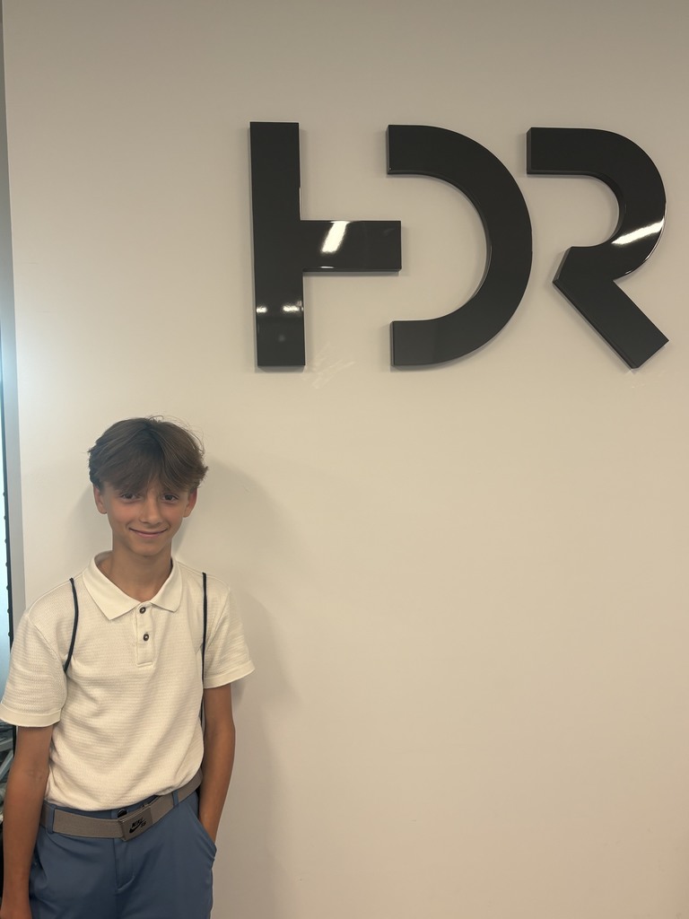 A young boy with light brown tousled hair stands by a white wall featuring large black 3D letters.  He smiles, wearing a white polo shirt, light blue pants, a grey Nike belt, and has backpack straps visible on his shoulders.