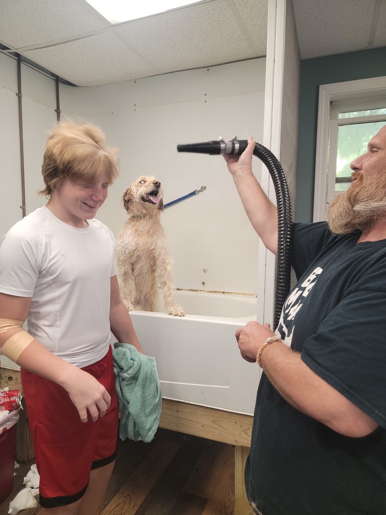 A bearded man holds a vacuum cleaner aimed at a young person with light, tousled hair who is smiling with eyes closed. The young person wears a white t-shirt, red shorts, and an arm bandage, holding a green towel. Behind them, a scruffy, wet dog sits happily with tongue out on a raised platform in what appears to be an indoor grooming area.
