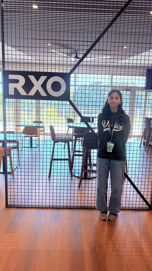 A young girl stands indoors in front of a glass door or partition with a grid pattern. She wears a black sweatshirt, light blue jeans, and brown shoes. Behind her is a room with blue and brown chairs and tables, large windows showing trees and buildings outside. She looks directly at the camera with long dark hair.