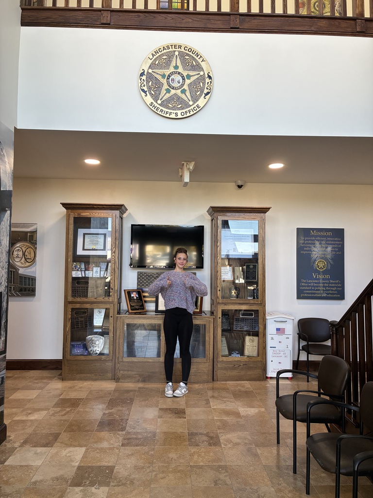 A woman stands indoors smiling and giving a thumbs-up with both hands in front of a display case and a wall emblem that reads "LANCASTER COUNTY SHERIFF'S OFFICE." The display contains awards and plaques. A sign nearby states the office's mission and vision. The woman wears a colorful sweater, black pants, and silver shoes. The room has wooden flooring and chairs.