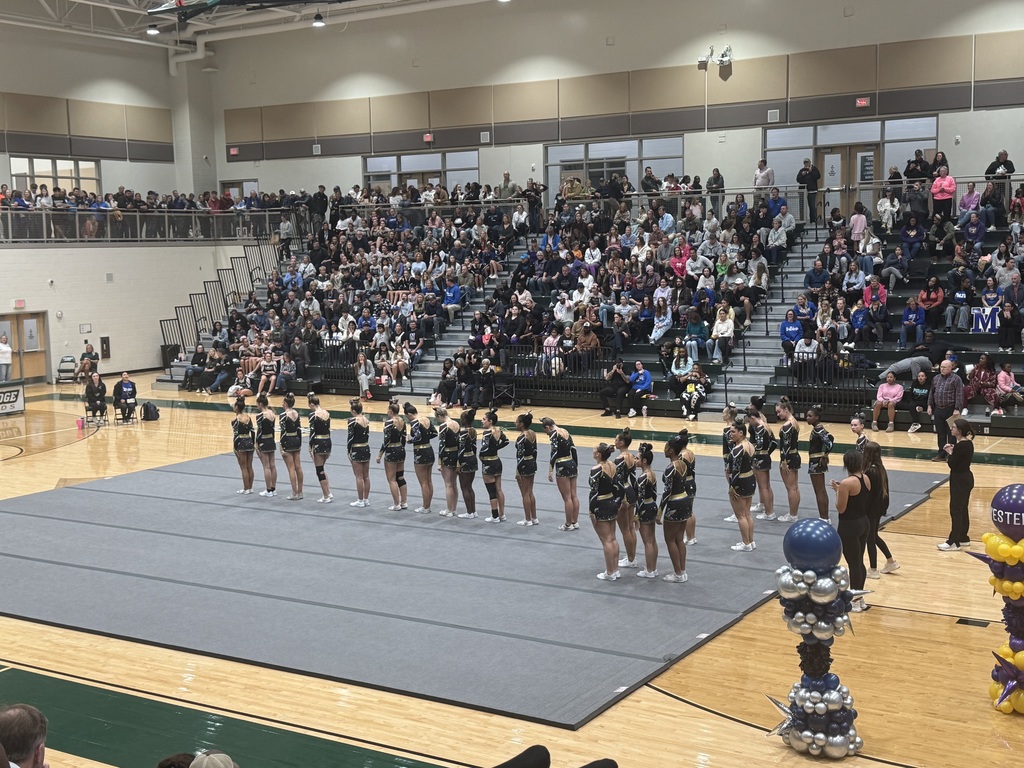 Indoor gymnasium filled with a large audience seated on tiered bleachers watching a cheerleading or dance team performance. The cheerleaders, dressed in black, gold, and white uniforms, stand in formation on a gray mat at center floor. Coaches or organizers stand nearby. Decorative balloon arrangements in black, gold, white, and purple are visible in the foreground. The background includes high ceilings with lights, walls with windows, and many spectators, indicating a well-attended event.