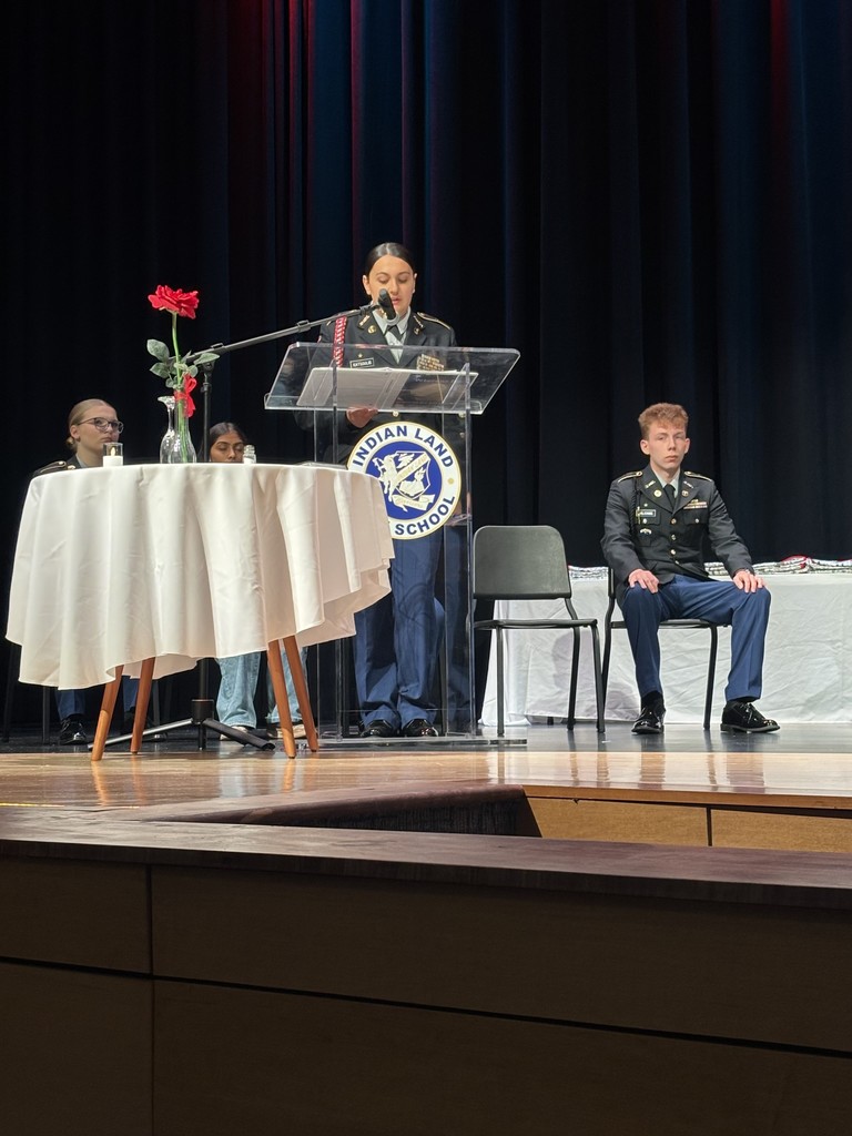 Four people in JROTC uniforms on a stage. One person stands at a clear podium with an "INDIAN LAND SCHOOL" emblem and is speaking. To the left is a table with a white tablecloth, a vase holding a red flower, and glasses and papers; a person is seated to the right. The backdrop is a black curtain; the event is a Veterans' Day ceremony.